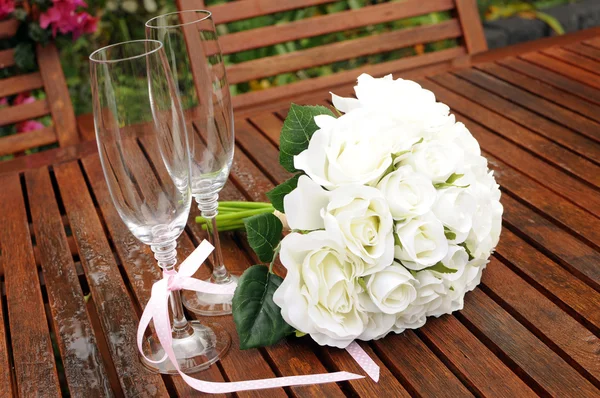 Wedding bridal bouquet of white roses with two champagne glasses with pink polka dot ribbon on outdoor garden table setting after rain. — Stock Photo, Image