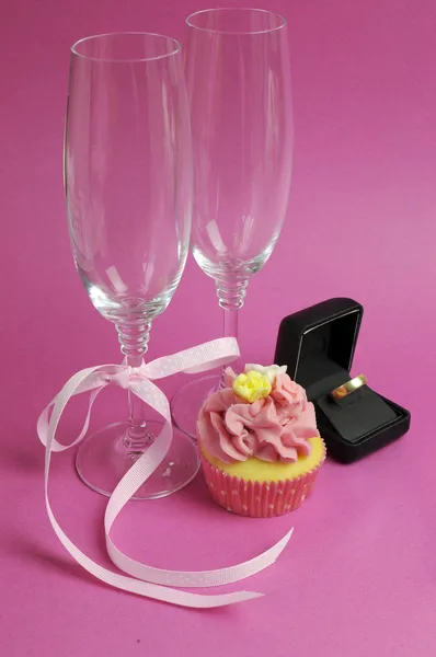Bröllop tema brud par champagne flöjt glasögon med rosa cupcake och vigselring i svart smyckeskrin mot en rosa bakgrund. vertikala. — Stockfoto