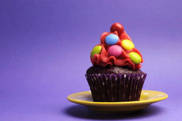 Doces brilhantes coberto cupcake na placa de bolinhas amarelas contra um fundo roxo — Fotografia de Stock