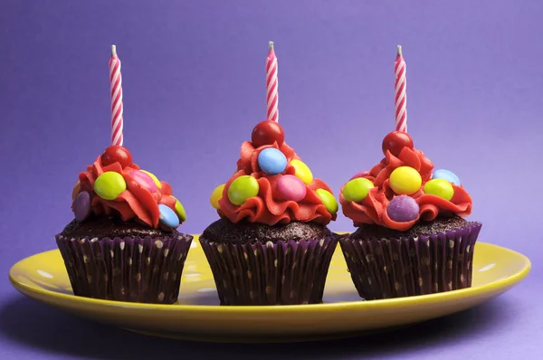 Drei bunte bonbonbezogene Cupcakes mit Geburtstagskerzen auf gelbem Teller vor violettem Hintergrund — Stockfoto