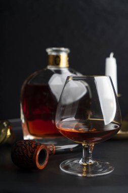 Bir bardak konyak ve tütün için bir pipo bir şişe konyak ve koyu bir yüzey üzerinde bir mum.