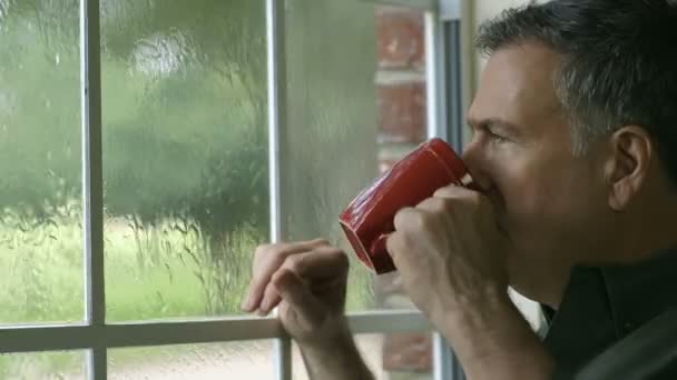 ein schöner, reifer Mann genießt eine Tasse Kaffee, während er einen Sommerregen durch sein Fenster sieht.