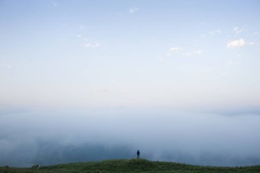 Minik İnsan, arka planda çimen ve bulutların bulunduğu dağlık alanda yürüyor. İnsan doğayla baş başa..