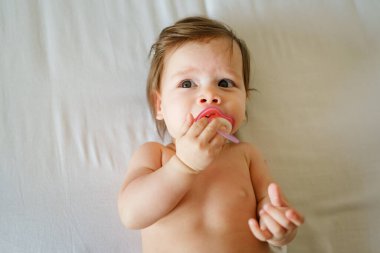 Beyaz, küçük bir kız bebek. Altı aylık. Yatakta çıplak, meme ucu emziği parlak.