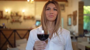 Güzel mutlu esmer beyaz kadın portresi. Elinde bir bardak kırmızı şarap içerken gülümsüyor. Restoranda kameraya bakıyor.