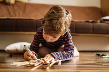 Evinde yerde oynayan küçük beyaz çocuk kağıt oyuncaklarından resim yapmak için boya kalemi kullanıyor. Yeni yetenekler geliştiriyor. Oyuncak çocuk büyüyen eğlenceli bir konsept.