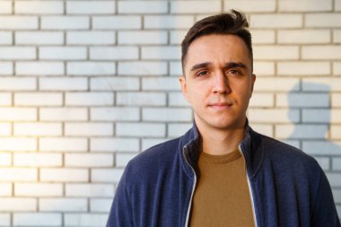 Beyaz tuğla duvarın önünde duran beyaz tenli genç bir adamın ön manzara portresi. Kameradaki boşluğa bakıyor.