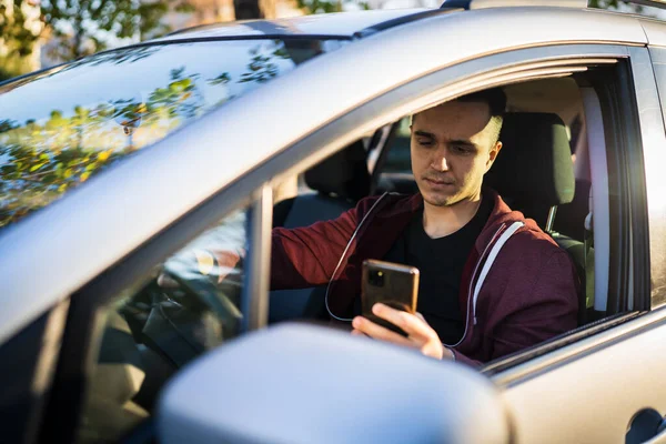 若い白人男性のフロントビュー車の運転や駐車場に座っている間に目的地のためのメッセージやナビゲーションアプリの携帯電話をチェック本物の人々の旅行のコンセプト — ストック写真