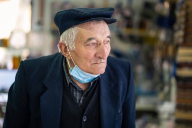 Son sınıf emeklisi, eski erkek portresi. Kapalı mekâna bakıyor. Çenesinde maske, başında şapka var.
