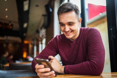 Kafe 'deki masada oturan beyaz bir gencin portresi. Elinde cep telefonu var. Mesajlar okuyor.