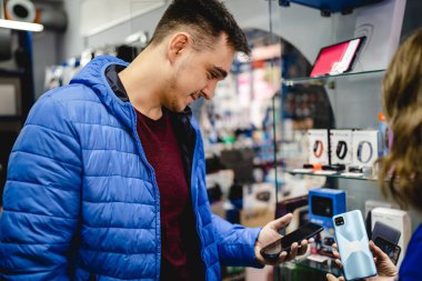 Elektronik mağazasında dikilen genç, beyaz bir adamın portresi. Cep telefonu görünümlü, mavi ceketli, gerçek insanlar uzay kopyaları yapıyor.