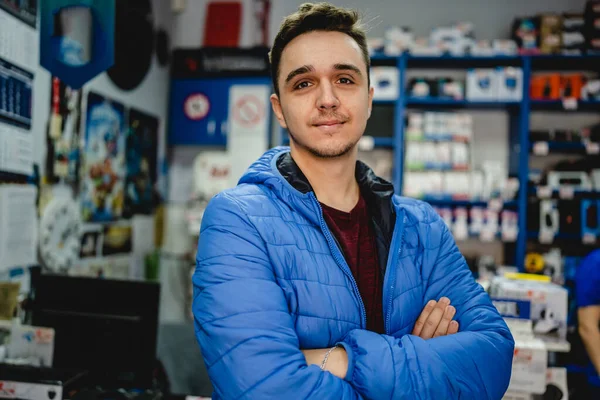 Front view portrait of one young adult caucasian man standing in the electronics store serious looking to the camera wearing blue jacket real people