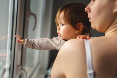 Beyaz kadın anne, küçük bebeğini kucağında tutarken pencerenin kenarında durup gerçek insanların sevgi ve aile kavramını birleştiren ebeveynlerine bakıyor.