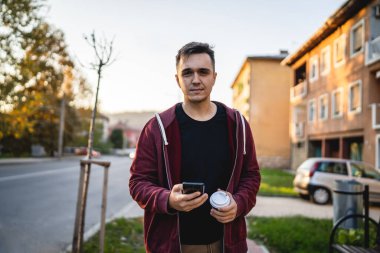 Sonbahar günü elinde cep telefonu ve bir fincan kahveyle şehrin önünde dikilen yetişkin beyaz bir adamın ön görüntüsü. Gerçek insanlar kameraya bakarak mekanı kopyalıyorlar.