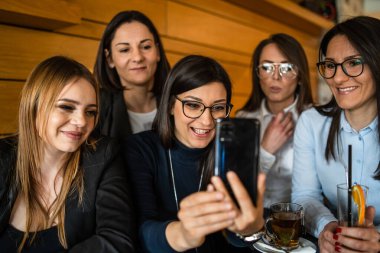 Kafe ya da restoranda bin yıllık arkadaşlarıyla oturmuş akıllı telefondan video görüşmesi yaparken ya da selfie çekerken cep telefonu tutan bir grup yetişkin beyaz kadının ön görüntüsü.