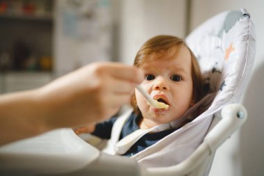 Kimliği belirsiz bir annenin sandalyesinde oturan küçük bir beyaz bebek evde plastik kaşıklı küçük yedi aylık bebeğe yemek verirken gerçek insanlar mekanı kopyalamayı önemsiyor.