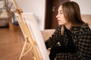 Beyaz tenli genç bir kadın sanatçı evinde, kanepede oturup resim yapıyor elinde boya fırçası ve yaratıcılık, profesyonel ya da hobi kavramları var.