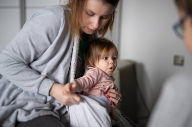 Küçük beyaz bir bebek giyiniyor. Genç bir kadın, küçük kızını kucağına alıp üstünü değiştiriyor. Gerçek insanlar, aile içi bakım ve ebeveynlik kavramı.