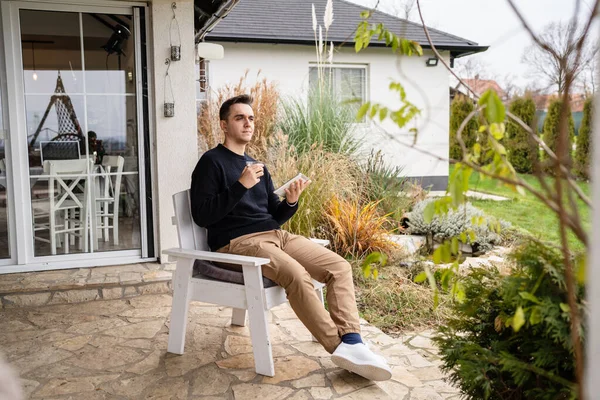 一位年轻的白人男子在屋前的椅子上坐着 手里拿着一杯咖啡和数码平板电脑 向旁边看去 — 图库照片