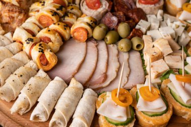 Peynir ve jambon rulolarını ahşap tabakta kapayın. Yemeye hazır çeşitli yemekler var. Aperatif masada.