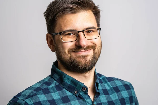 30 yaşında, sakallı ve gözlüklü beyaz bir adamın portresi. Beyaz bir duvarın önünde kameraya bakıyor. Sırıtıyor.