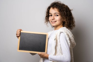 On yaşında beyaz bir kız, kıvırcık saçlı, önden görünüşlü. Beyaz arka plan önünde duruyor. Metin fotokopisi için kameraya bakıyor.