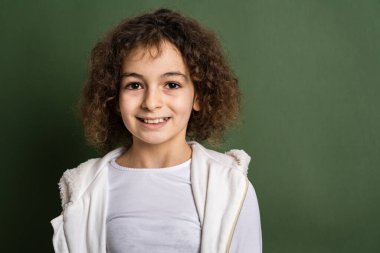 On yaşında, kıvırcık saçlı, yeşil arka planda dikilip kameraya bakıp gülümseyen mutlu ve neşeli fotokopi çeken bir beyaz kız.