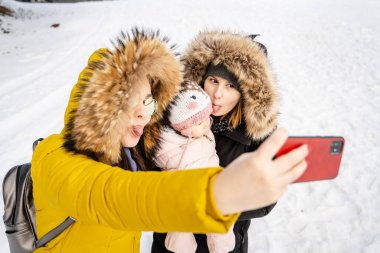 İki beyaz kadın ve bebek, küçük çocuğu ve arkadaşıyla selfie çekiyor. Ya da teyzesi fotoğraf çekiyor. Ya da kış günü dışarıda kar yağarken cep telefonuyla arama yapıyor.