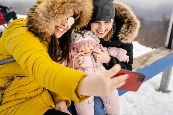 Two caucasian women and baby girl taking selfies mother with her small child and her friend or aunt taking photos or making a video call using mobile phone in winter day in nature with snow outdoor