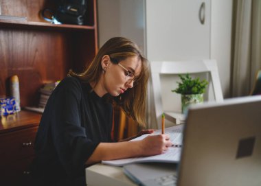 One young caucasian woman female student sitting at home writing in her notebook holding pencil while study preparing for exam learning and education concept real people copy space side view