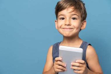 Dört yaşında, mavi arka plan stüdyosunun önünde dikilip dijital tablet kullanarak kameraya gülümseyen küçük beyaz bir çocuğun ön görüntüsü.