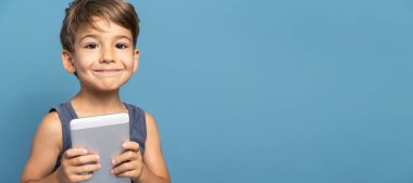 Dört yaşında, mavi arka plan stüdyosunun önünde dikilip dijital tablet kullanarak kameraya gülümseyen küçük beyaz bir çocuğun ön görüntüsü.