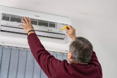 Beyaz tenli yaşlı bir adamın evdeki klimayı tamir edişinin arka görüntüsü.