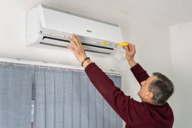 Beyaz tenli yaşlı bir adamın evdeki klimayı tamir edişinin arka görüntüsü.