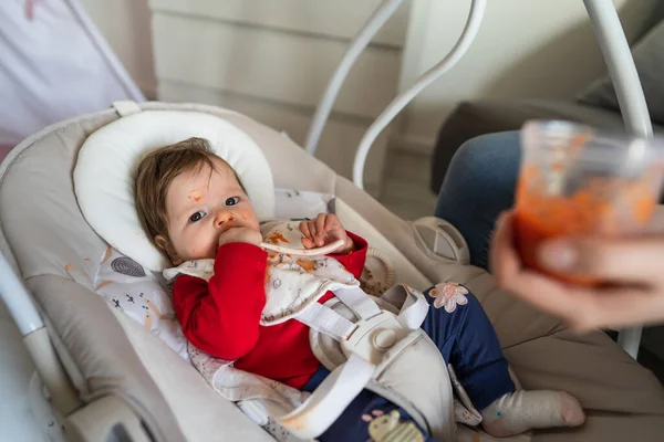 First meal problems concept small caucasian baby five months old refusing to eat spitting carrot mash puree mess on her face dirty messy hand of unknown mother holding the spoon while feeding