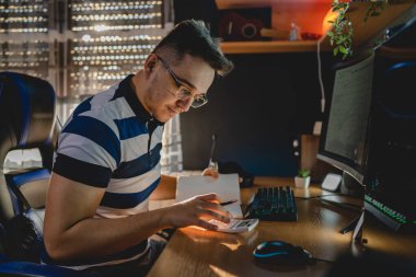 Genç bir yetişkin beyaz erkek, masa ve bilgisayar başında otururken kripto para kazanma işi ve ticaretine yaptığı yatırımı hesaplıyor.
