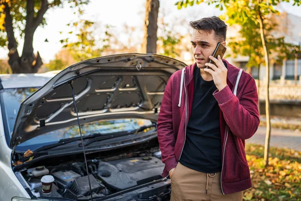 秋の日に道路脇援助の概念の助けを求める電話牽引サービスを保持していないオープンフードと壊れたエンジンを持つ彼の車のそばに立っている1人の若い大人の白人男性 — ストック写真