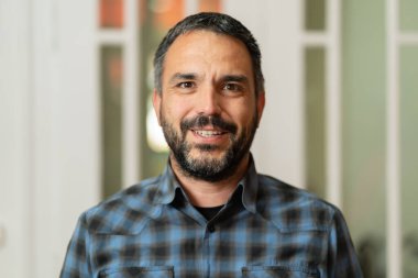 Sakallı, kısa saçlı, mutlu beyaz bir adamın resmi. Ofiste ya da evde tişört giymiş kameraya bakıyor.