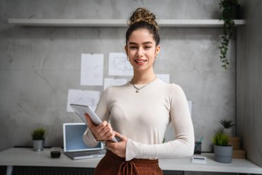 Genç, beyaz, mutlu, başarılı bir bayan esmer iş yerinde dikiliyor. Elinde dijital tablet beliyle ön tarafta fotokopi odası var. Kameraya bakıp gülümsüyor.
