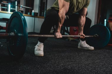 Yetişkin, beyaz, erkek, vücut geliştirme eğitimi almış bir adam. Spor salonunda dikilirken ağırlık kaldırıyor. Üzerinde koyu renk bir fotoğraf var.