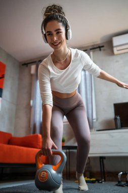 Genç bir kadın, odasında spor yapıyor. Elinde kettlebell ağırlığı ve kafasında kulaklıklarla duruyor. Modern kıvırcık esmer ev antremanı.