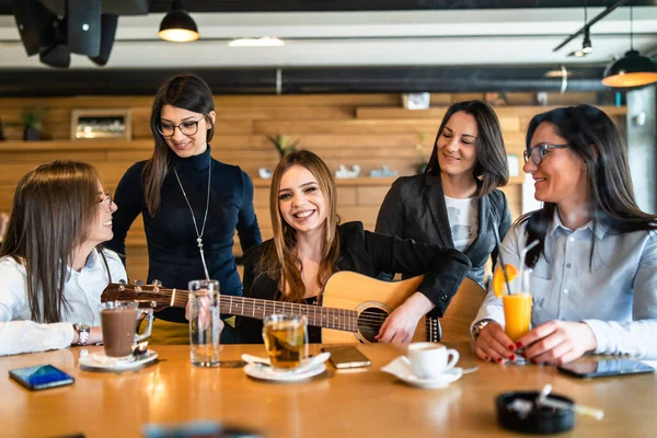 Young millennial women sitting by the table at cafe or restaurant singing and smiling having fun with guitar - Beautiful caucasian women having a good time while playing guitar - friendship concept