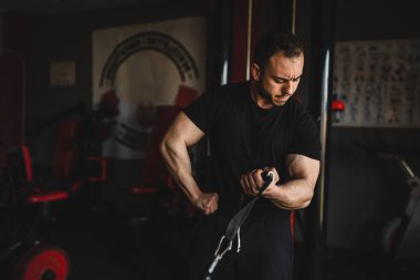 Yetişkin, beyaz, erkek, vücut geliştiren bir adam spor salonundaki kablo TV makinesinde silah eğitimi alıyor. Ağırlık tutuyor, siyah tişört giyiyor. Siyah tişört giyiyor.
