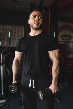 Salonda elinde dambıllarla duran yetişkin bir vücut geliştirici.