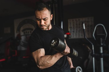 Yetişkin, beyaz, erkek, vücut geliştiren, kollarını çalıştıran, spor salonunda siyah tişörtlü, siyah fotograflı, kaslarını esneten bir adam.