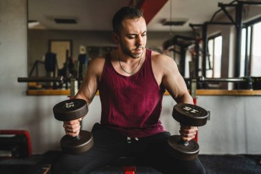 Yetişkin, beyaz, erkek, vücut geliştiren, spor salonunda oturmuş gerçek insanların giydiği gömleği giyerken kol kaslarını halterle esneten bir adam.