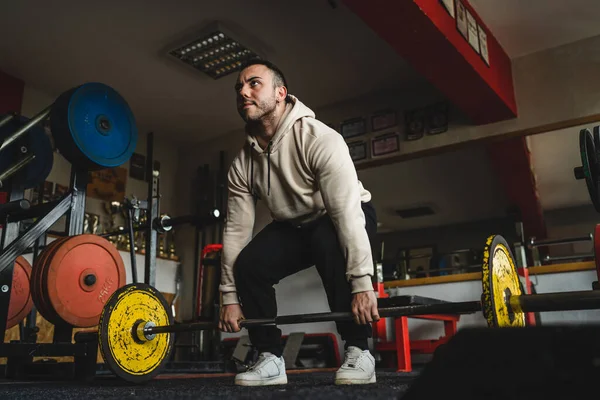 一位年轻的白人男性健美运动员穿着连帽衫站在体育馆里 一边用重物和理发店的重物做着举重运动 一边训练着自己的健美健美身体 — 图库照片