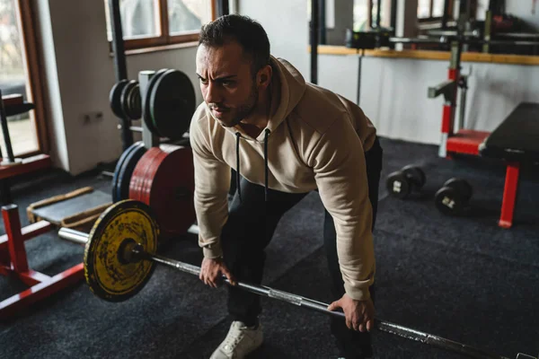 One man young adult caucasian male bodybuilder training back doing deadlift with barbel and weights while standing in the gym wearing hoodie real people copy space side view full length