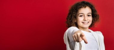 On yaşında beyaz bir kız, kıvırcık saçlı ön plan portresi kırmızı arka planda dikilip kameraya bakarak mutlu ve neşeli fotokopi çekerken gülümsüyor.