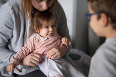Küçük beyaz bir bebek giyiniyor. Genç bir kadın, küçük kızını kucağında tutuyor. Onu evde giydirip, erkek kardeşiyle kıyafetlerini değiştiriyor.